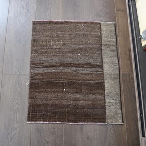 Puede incluir: Una alfombra rectangular tejida a mano, con una combinación de colores marrón y beige. La alfombra tiene una apariencia texturizada y un borde de un tono más claro. Está colocada sobre un suelo de madera.