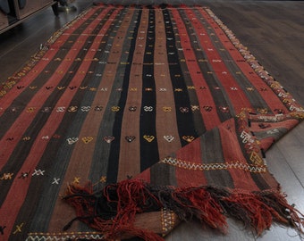 May include: A long, narrow, handwoven rug with a striped pattern in shades of red, black, and brown. The rug has a fringe on both ends.