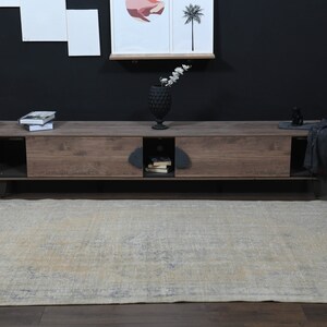 May include: A light beige rug with a faded floral pattern. The rug is placed in front of a wooden entertainment center with black metal legs. The entertainment center has two drawers and an open shelf.
