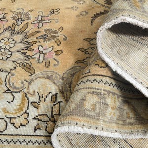 May include: A vintage-style area rug with a floral pattern in beige, cream, and muted pink tones. The rug's design features detailed floral motifs and a rolled-up section revealing the underside, showcasing its construction and fringe.