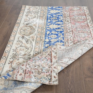 Könnte beinhalten: Ein rechteckiger, abgenutzter Teppich in Creme, Blau und Rosa. Der Teppich hat florale und geometrische Muster. Er ist teilweise aufgerollt, wodurch die Unterseite sichtbar wird, und liegt auf einem dunklen Holzboden. Maße in Zentimetern.