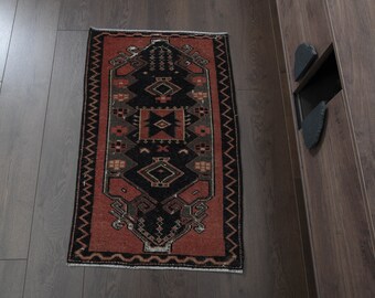 May include: A red, black, and green patterned rug with a geometric design. The rug has a fringe border and is placed on a wooden floor.
