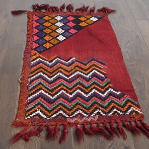 May include: A red, handwoven rug with a geometric pattern of zigzags and diamonds in shades of orange, green, white, and black. The rug has a fringe border.