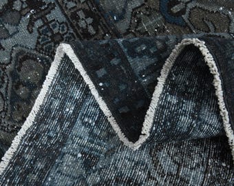May include: A folded area rug featuring a dark blue and charcoal gray color palette with a complex, repeating geometric and floral pattern. The rug has a visible white fringe along the edge where the fabric is folded, revealing the woven texture.