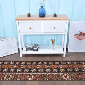 Op de afbeelding: Een bruine en zwarte loper met geometrische patronen. De loper ligt voor een witte consoletafel met twee laden en een onderplank. De tafel staat tegen een lichtblauwe muur.