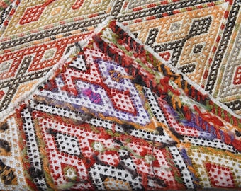 May include: A textile featuring a complex geometric pattern with diamond shapes in shades of red, orange, yellow, black, white, and purple. The fabric displays intricate cross-stitch embroidery with visible thread ends and a folded corner edge.