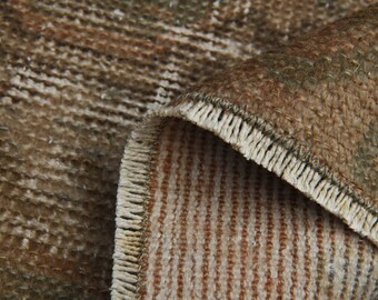 May include: Close-up of a section of a rug with a distressed, vintage appearance. The rug features a mix of brown, beige, and green tones, with visible texture and a fringed edge. The design includes vertical stripes and faded patterns.