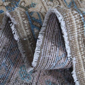 Könnte beinhalten: Nahaufnahme eines gefalteten, abgenutzten Teppichs mit verblichenem, Vintage-Aussehen. Der Teppich weist ein komplexes Muster in Beige-, Braun- und Blautönen auf. Die Kanten sind ausgefranst, was die antike Ästhetik unterstreicht. Die Textur des Teppichs ist sichtbar.