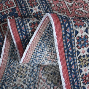 Könnte beinhalten: Nahaufnahme eines gefalteten, gemusterten Teppichs mit Used-Look. Der Teppich weist ein sich wiederholendes geometrisches Design in Rot-, Blau- und Beigetönen auf. Die Kanten haben einen Fransenrand.
