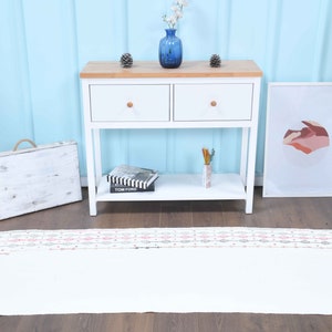 May include: White runner rug with a geometric pattern in shades of pink, gray, and white. The rug is placed in front of a white console table with two drawers and a lower shelf. A framed print with an abstract design hangs on the wall behind the table.