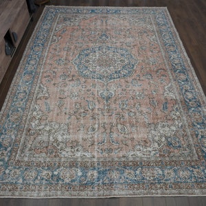 Op de afbeelding: Een vintage-stijl tapijt met een vervaagd roze en blauw bloemenpatroon. Het tapijt heeft een verweerde uitstraling en een versleten rand.