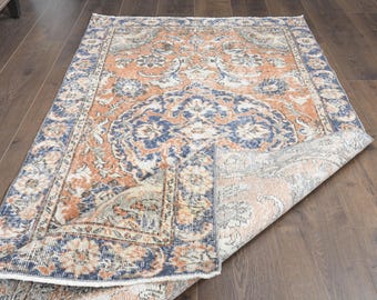 May include: A vintage-style rug with a floral pattern in shades of blue, orange, and cream. The rug is folded in half, revealing the back side, which is a lighter cream color.