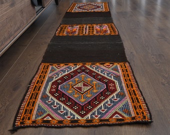 May include: A long, narrow rug with a black background and colorful geometric patterns. The rug is made of wool and has a fringed edge.