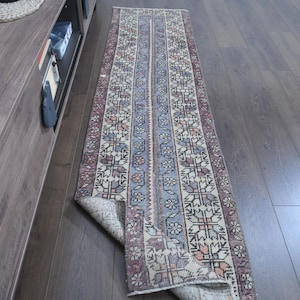 Könnte beinhalten: Ein rechteckiger Läuferteppich mit einem floralen und geometrischen Muster in Blau-, Creme- und Mauve-Tönen. Der Teppich liegt auf einem dunklen Holzboden, wobei ein Teil des Teppichs umgeschlagen ist.