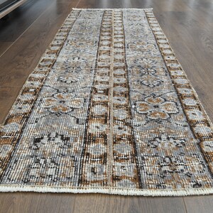 May include: A rectangular, vintage-style rug with a distressed pattern. The rug features a central design of vertical stripes in grey, brown, and cream tones, with floral motifs. The rug is placed on a dark wood floor.
