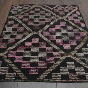 Peut inclure: Un tapis rectangulaire noir avec un motif géométrique. Le motif présente des carrés et des lignes diagonales dans des tons de rose, beige et blanc. Le tapis est posé sur un sol en bois foncé.