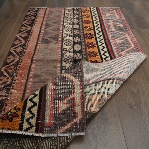 Puede incluir: Una alfombra vintage con un patrón geométrico colorido en tonos marrones, naranjas y rojos. La alfombra está doblada por la mitad, revelando el reverso, que es de color blanco roto.