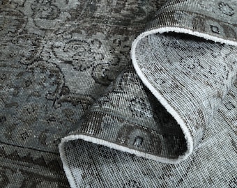 May include: A rolled-up, distressed-look rug with a faded, muted color palette. The rug features a detailed, traditional pattern in shades of gray, charcoal, and hints of blue. The edges are finished with a white fringe, adding a touch of contrast.