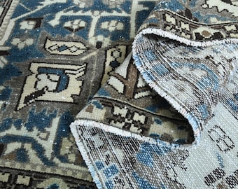 May include: An antique rug with a detailed pattern in shades of blue, beige, and brown. The rug is partially rolled up, revealing the underside with a fringe edge. The design features geometric shapes and floral motifs.