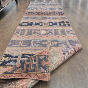 Pode incluir: Um tapete corredor retangular com um padrão geométrico em tons de azul, pêssego e bege. O tapete está enrolado em uma extremidade, revelando a parte inferior. O design apresenta formas e linhas repetidas.