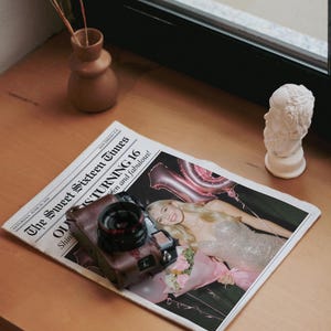 May include: A brown leather camera rests on a newspaper titled "The Sweet Sixteen Times." The newspaper features a photo of a woman in a sparkly dress, with the headline "STURNING 16." A small bust and a vase with dried flowers are also on the wooden surface.
