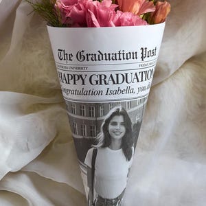 K&ouml;nnte beinhalten: Ein Strau&szlig; rosa Blumen, verpackt in einem Zeitungskegel mit der &Uuml;berschrift "The Graduation Post" und dem Text "HAPPY GRADUATION Congratulation Isabella, you did it!" und "Class of..."