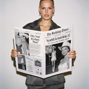 May include: A woman in a gray blazer and skirt holds a newspaper titled "The Birthday Times." The newspaper features the headline "Scarlett is turning 25!" and party-themed images. The woman has her hair up and is wearing hoop earrings.