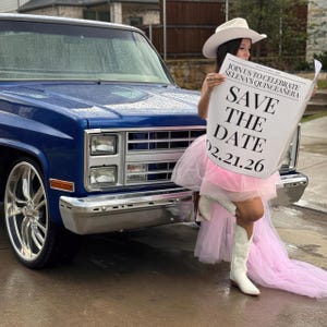 May include: A blue vintage pickup truck with chrome accents. A person is holding a newspaper that says "Join us to celebrate Selena's Quinceanera" and "Save the Date 02.21.26". The person is wearing a white cowboy hat, pink dress, and white cowboy boots.