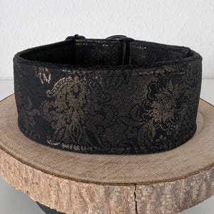 Könnte beinhalten: Ein schwarzes Hundehalsband mit goldenem Blumenmuster. Das Halsband besteht aus einem dicken Stoff und hat eine schwarze Kunststoffschnalle. Das Halsband wird auf einer runden Holzscheibe präsentiert.