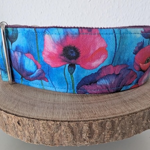 Könnte beinhalten: Ein breites Hundehalsband mit einem floralen Muster aus rosa und lila Mohnblumen auf blauem Hintergrund. Das Halsband hat eine silberne Metallschnalle und wird auf einer Holzscheibe präsentiert.