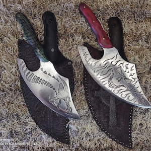 Puede incluir: Dos cuchillos decorativos con vainas de cuero. El cuchillo de la izquierda tiene un mango verde y un diseño de dragón en la hoja. El cuchillo de la derecha tiene un mango rojo y un diseño de calavera con llamas en la hoja.