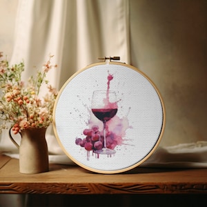 Puede incluir: Un aro de bordado blanco con una copa de vino tinto y uvas en un diseño de estilo acuarela. La copa está llena de vino tinto y hay salpicaduras de vino tinto alrededor de la copa.