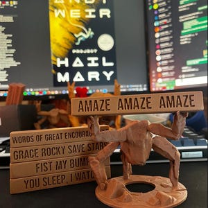 May include: A brown 3D-printed figure with multiple limbs holding a rectangular sign that reads AMAZE AMAZE AMAZE. Beside it, a stack of four brown rectangular blocks features the text WORDS OF GREAT ENCOURAGEMENT, GRACE ROCKY SAVE STARS, FIST MY BUMP, and YOU SLEEP, I WATCH.