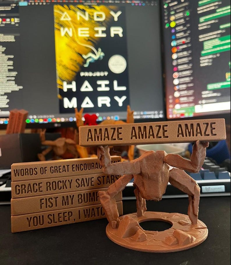 May include: A brown 3D-printed figure with multiple limbs holding a rectangular sign that reads AMAZE AMAZE AMAZE. Beside it, a stack of four brown rectangular blocks features the text WORDS OF GREAT ENCOURAGEMENT, GRACE ROCKY SAVE STARS, FIST MY BUMP, and YOU SLEEP, I WATCH.