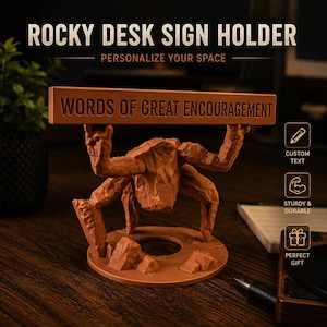 Puede incluir: Un soporte de escritorio marr&oacute;n con textura de roca que muestra una figura con los brazos levantados sosteniendo una barra rectangular horizontal. La barra tiene grabado el texto WORDS OF GREAT ENCOURAGEMENT. La base es circular.
