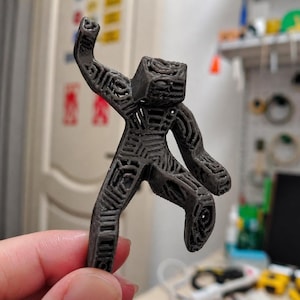 May include: A dark gray, 3D-printed figurine of a person in a dynamic pose, with one arm raised and a leg bent. The figure has a geometric, open-frame design, showcasing the internal structure. The figurine is held in a hand.