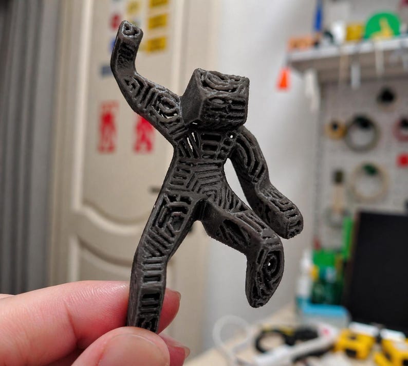 May include: A dark gray, 3D-printed figurine of a person in a dynamic pose, with one arm raised and a leg bent. The figure has a geometric, open-frame design, showcasing the internal structure. The figurine is held in a hand.