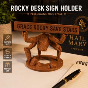 Puede incluir: Un soporte de letrero de escritorio marr&oacute;n texturizado con una criatura de varias patas sobre una base circular con rocas, que sostiene un letrero rectangular horizontal con el texto GRACE ROCKY SAVE STARS en letras may&uacute;sculas.
