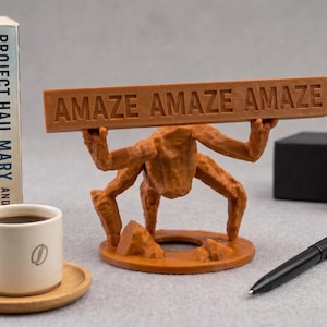 Puede incluir: Una figura de criatura marr&oacute;n de m&uacute;ltiples patas sobre una base circular, sosteniendo una barra rectangular horizontal sobre su cabeza. La barra presenta el texto en relieve "AMAZE AMAZE AMAZE" en may&uacute;sculas. La figura tiene una superficie texturizada.