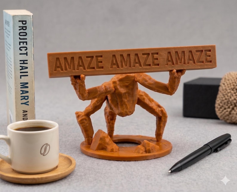 Puede incluir: Una figura de criatura marr&oacute;n de m&uacute;ltiples patas sobre una base circular, sosteniendo una barra rectangular horizontal sobre su cabeza. La barra presenta el texto en relieve "AMAZE AMAZE AMAZE" en may&uacute;sculas. La figura tiene una superficie texturizada.
