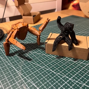 May include: Two 3D-printed figurines. One is a brown, insect-like creature with articulated limbs. The other is a black, abstract figure with a patterned design, standing on a small cardboard box. The figures are on a green cutting mat.