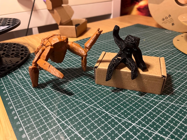 May include: Two 3D-printed figurines. One is a brown, insect-like creature with articulated limbs. The other is a black, abstract figure with a patterned design, standing on a small cardboard box. The figures are on a green cutting mat.