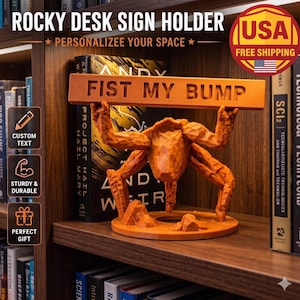 May include: An orange desk sign holder featuring a stylized figure with a textured, carved appearance holding a horizontal bar above its head. The bar is embossed with the text FIST MY BUMP in capital letters. The figure stands on a circular base.