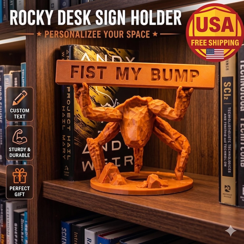 May include: An orange desk sign holder featuring a stylized figure with a textured, carved appearance holding a horizontal bar above its head. The bar is embossed with the text FIST MY BUMP in capital letters. The figure stands on a circular base.