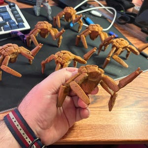 May include: A collection of brown, crab-like figurines with detailed textures. The figurines are arranged on a dark surface, with one held in a hand. The crab figurines have multiple legs and a segmented body, suggesting a marine or fantasy theme.