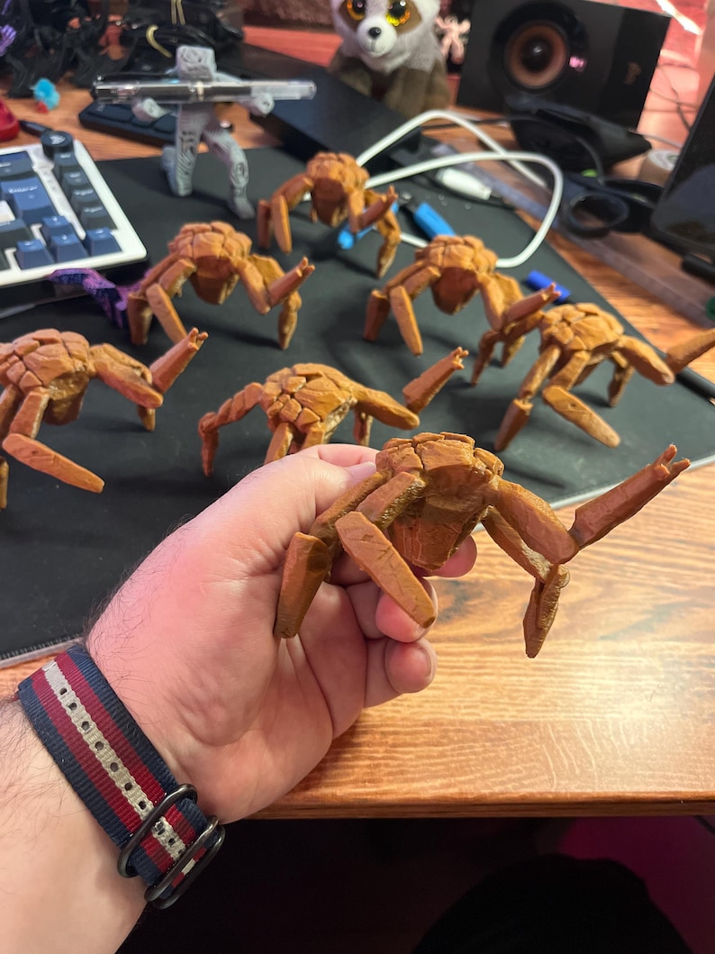 May include: A collection of brown, crab-like figurines with detailed textures. The figurines are arranged on a dark surface, with one held in a hand. The crab figurines have multiple legs and a segmented body, suggesting a marine or fantasy theme.