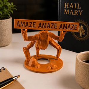 Puede incluir: Una figura de criatura naranja de m&uacute;ltiples patas con textura, sobre una base circular, sosteniendo un letrero rectangular horizontal sobre su cabeza. El letrero presenta el texto en relieve "AMAZE AMAZE AMAZE" en una fuente negrita.