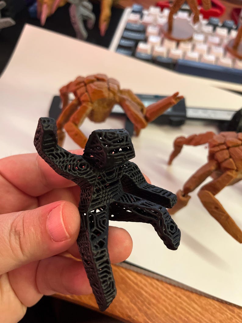 May include: A black, 3D-printed figurine of a person with outstretched arms, held in a hand. The figurine has a detailed, open lattice design. Other similar figurines in shades of brown and gray are visible in the background.
