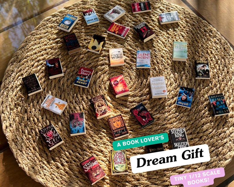 May include: A collection of tiny 1/12 scale books, including titles like "Doctor Sleep" and "Atomic Habits," arranged on a woven mat. The image includes text that reads "A Book Lover's Dream Gift."