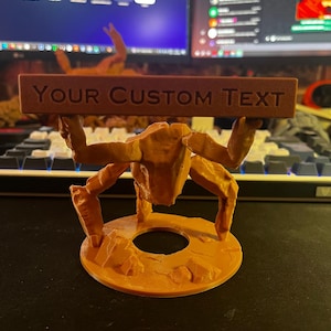 Puede incluir: Una figura de pl&aacute;stico naranja impresa en 3D con textura rocosa, de pie sobre una base circular y sosteniendo una barra horizontal rectangular sobre su cabeza. La barra presenta el texto en relieve "YOUR CUSTOM TEXT".